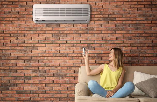 Woman operating air conditioner while sitting on sofa at home - Fotó, kép