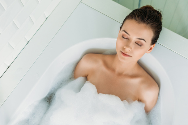 top view of beautiful young woman with closed eyes relaxing in bathtub - Фото, изображение