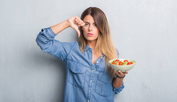 Молода доросла жінка над сірою гранжевою стіною їсть здоровий томатний салат з розлюченим обличчям, негативний знак, що показує нелюбов до великих пальців вниз, концепція відторгнення
 - Фото, зображення