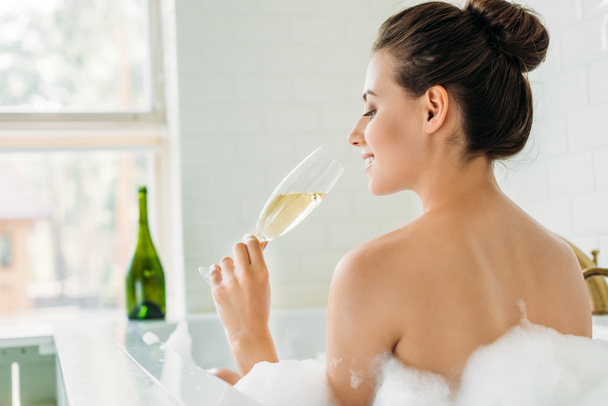 back view of smiling young woman drinking champagne in bathtub with foam - Foto, Imagen