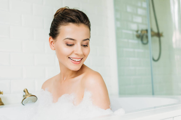 beautiful smiling young woman looking down at foam in bathtub  - Фото, изображение