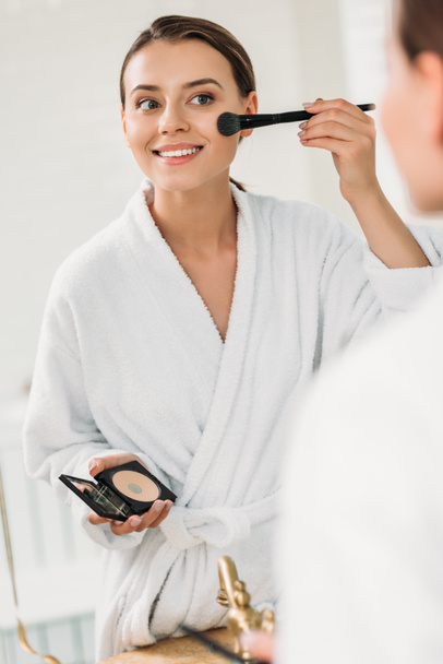 sorrindo jovem mulher em roupão de banho olhando para o espelho e aplicando maquiagem
 - Foto, Imagem