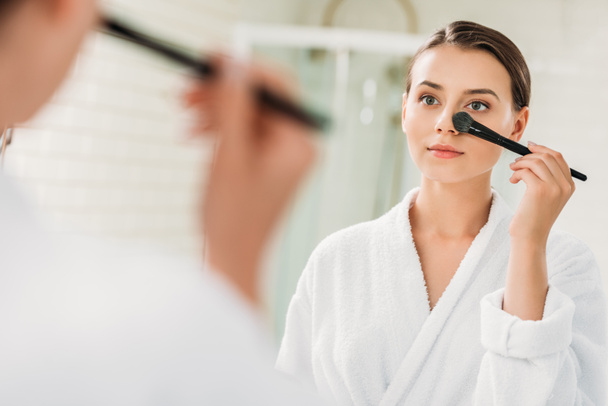 selective focus of beautiful brunette girl in bathrobe looking at mirror and applying makeup - Фото, изображение