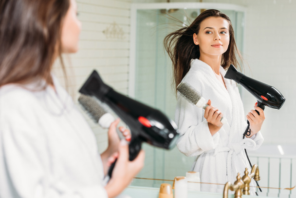 красива молода жінка тримає щітку для волосся і сушить волосся у дзеркалі у ванній
 - Фото, зображення