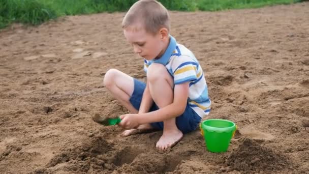 Ein kleiner Junge gräbt mit einer Schaufel eine Grube. Kind spielt am Strand - Filmmaterial, Video
