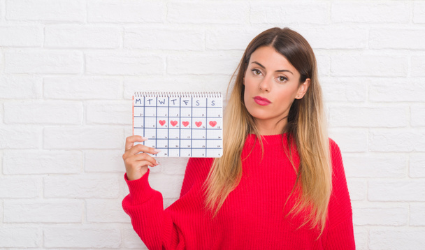 Mladé dospělé ženy nad bílé zdi drží menstruační kalendář s výrazem věří na inteligentní tvář myslí vážně - Fotografie, Obrázek