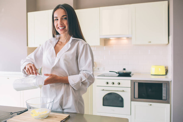 Positive woman stands in kitchen and smiles. She is pouring milk into big glass bowl. She wears white shirt. - Fotoğraf, Görsel