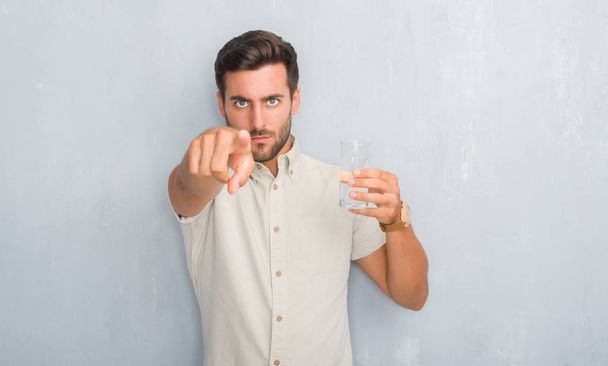 Beau jeune homme sur le mur gris grunge boire verre d'eau pointant du doigt vers la caméra et à vous, signe de la main, geste positif et confiant de l'avant
 - Photo, image