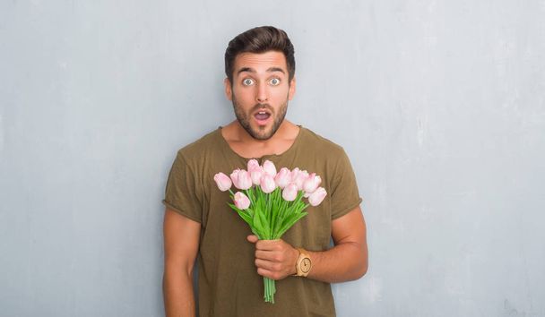 Beau jeune homme sur le mur gris grunge tenant bouquet de fleurs effrayé sous le choc avec un visage surprise, effrayé et excité par l'expression de la peur
 - Photo, image