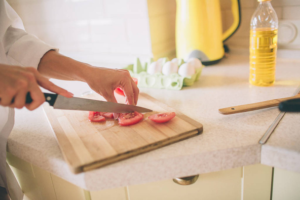 Close up of girls hands holding knife and cutting tomato into small pieces. She does it on wood board. Girl is cooking breakfast. - Foto, Bild