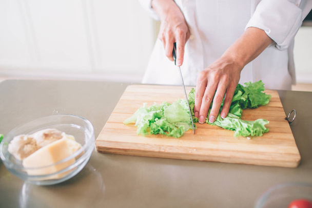 Cut view of girls hands cutting lettuce on wood board with knife. There is a bowl with boiled chicken and cheese besides it. - Foto, imagen