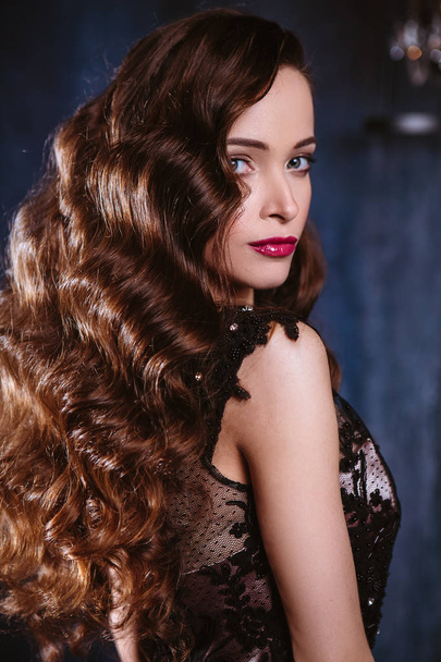 Young woman with long curly hair and makeup in evening long luxury dress, posing in a dark interior room. fashion beauty portrait - Foto, immagini