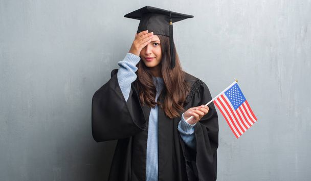 Jeune femme brune sur le mur gris grunge portant uniforme diplômé tenant drapeau de l'Amérique stressé avec la main sur la tête, choqué par la honte et le visage surprise, en colère et frustré. Peur et contrariété par erreur
. - Photo, image