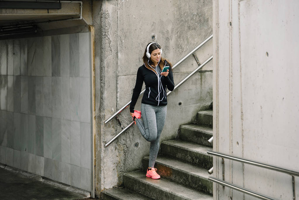 Sporty woman with headphones stretching legs for warming up before running in the city. Female athlete exercising and listening music with smart phone. - Foto, Bild