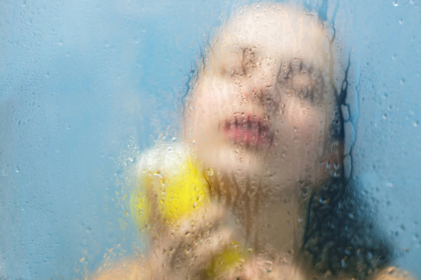 Relax in bathroom. Wet female keeps eyes closed, holds bath sponge, takes hot shower, wants to be clean and fresh, feels relaxation in shower cabine. Blurred sweat background. Wellness concept - Valokuva, kuva