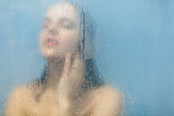 Unfocused silhouette of naked female model takes shower in cabine feels relaxed and takes care of her body, woman washes behind weeping glass shower door. Taking bath. Skin care concept - 写真・画像