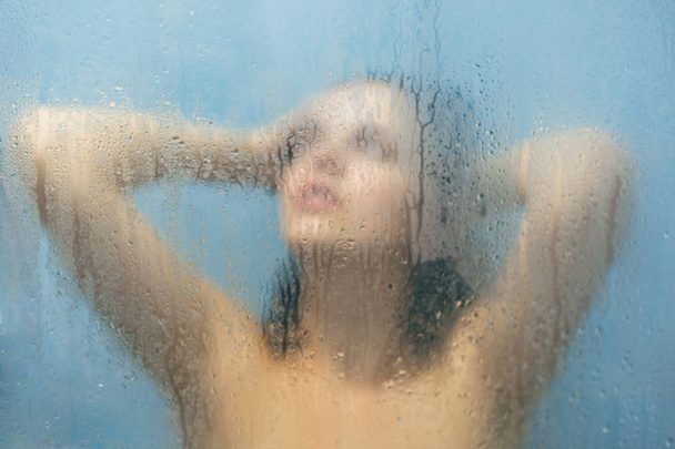 View of beautiful relaxed female with perfect naked body, takes shower, poses against blurrred glass sweat background, looks dreamy upwards, thinks and ponders about something. Wellness concept - Photo, Image