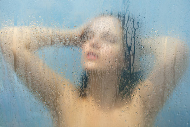 Female model washes in douche after stressful day, feels relief, poses naked in shower cabine. Unfocused woman`s silhouette in douche against blurred background with water drops. Care and beauty - Zdjęcie, obraz