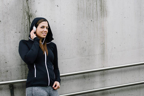 Urban fitness woman taking a workout rest for relaxing and listening music with headphones in city street. - Foto, Imagen