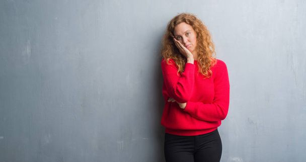 Gri grunge duvar kırmızı kazaklı düşünme seyir giyen genç Kızıl saçlı kadın yorgun ve sıkılmış çapraz silah depresyon sorunları ile. - Fotoğraf, Görsel