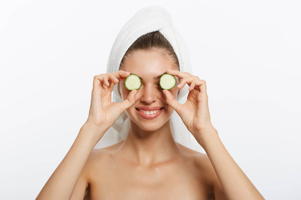Close up beauty portrait of a happy beautiful half naked woman with a towel wrapped around her head holding cucumber slices at her face isolated over white background - Foto, Bild