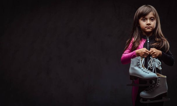 Μοναχικό κοριτσάκι ντυμένη με αθλητικά κατέχει παγοπέδιλα. Απομονωμένη σε ένα σκοτεινό φόντο με υφή. - Φωτογραφία, εικόνα