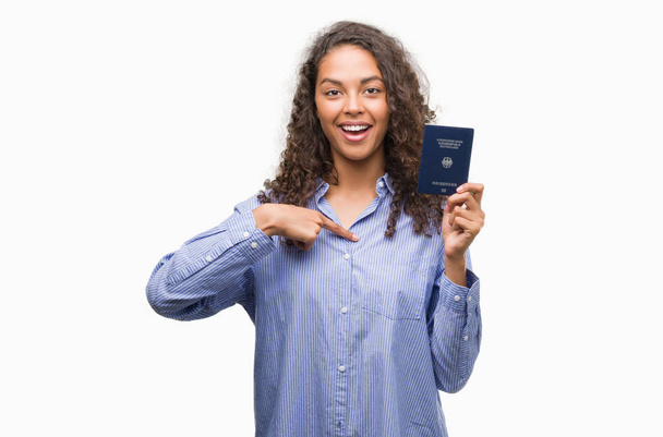 Νεαρή γυναίκα Ισπανόφωνος κατέχουν διαβατήριο της Γερμανίας με δάχτυλο που δείχνει πρόσωπο έκπληξη για τον εαυτό του - Φωτογραφία, εικόνα