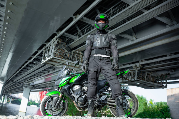 μοτοσικλετιστής σε ένα κράνος και προστατευτικό ρουχισμό που στέκεται κάτω από τη γέφυρα - Φωτογραφία, εικόνα