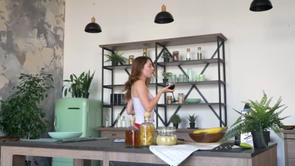 Attractive young woman dancing with glass of wine and smiling, modern kitchen in background - Video, Çekim