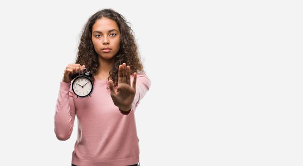 Young hispanic woman holding alarm clock with open hand doing stop sign with serious and confident expression, defense gesture - Фото, изображение