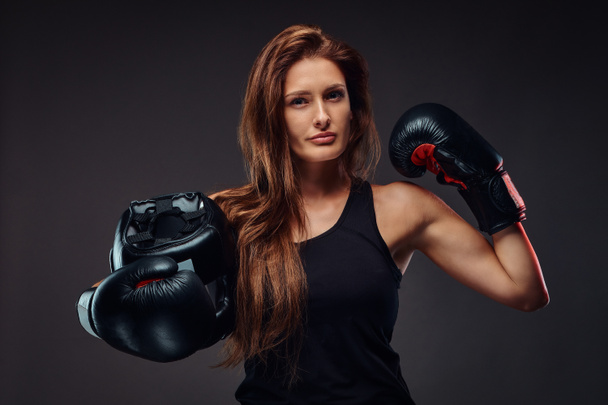 Mujer deportiva vestida con ropa deportiva con guantes de boxeo sostiene casco protector. Aislado sobre un fondo oscuro
. - Foto, Imagen