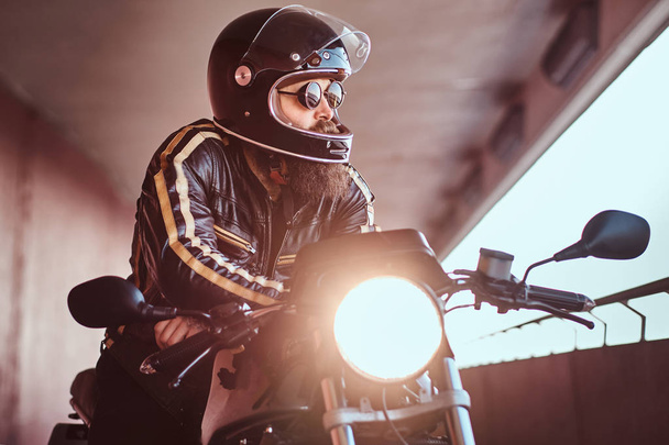 Close-up portrait of a brutal bearded biker in helmet and sunglasses dressed in a black leather jacket sitting on a retro motorcycle with included headlight. - Photo, Image