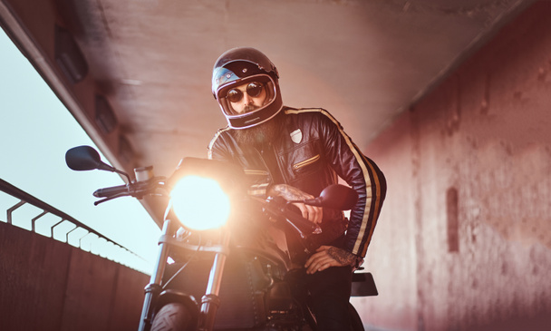 Retrato de cerca de un brutal motorista barbudo con casco y gafas de sol vestido con una chaqueta de cuero negro sentado en una motocicleta retro con faros incluidos
. - Foto, imagen