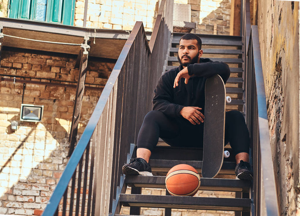 Close-up portrait of an African-American bearded hipster guy dressed in a black hoodie and sports shorts holds a skateboard while sitting on stairs in a ghetto. - Фото, изображение