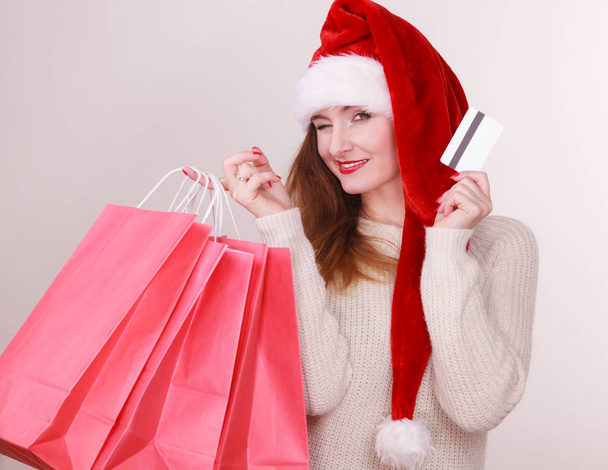 Noel zamanı. Holding kırmızı alışveriş torbaları ve kredi kartı, santa claus şapka giyen genç latin kadın hediyeler satın alma - Fotoğraf, Görsel