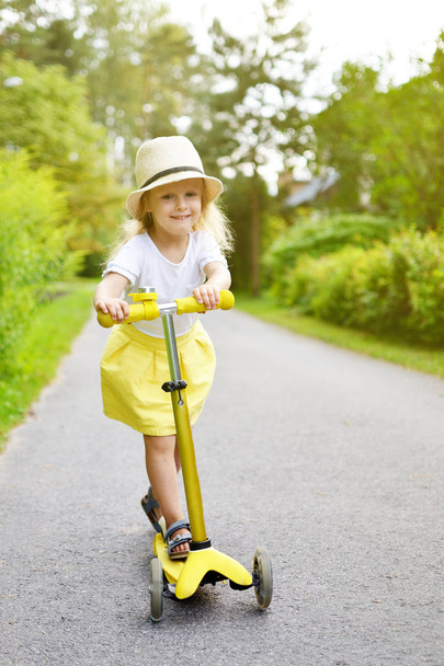 Gelukkig weinig gele rok wit Meisjesshirt rijden kick scooter buiten, het dragen van roze veiligheidshelm, leren evenwicht, zomertijd, zonlicht - Foto, afbeelding