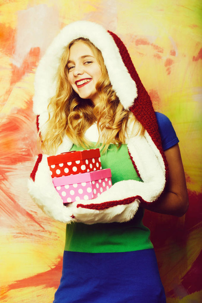 Happy pretty girl or cute woman with blond curly hair in red santa fur hood, knitted scarf and mittens smiles with gift boxes on pink and yellow wall on abstract background - Photo, Image