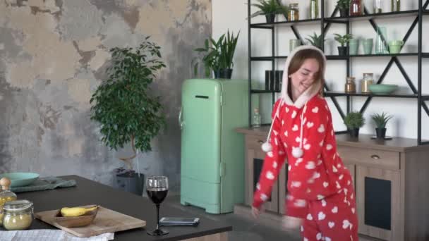 Happy young woman in pajamas dancing and smiling, wearing fancy hood, cheerful and funny, modern kitchen with green fridge - Imágenes, Vídeo