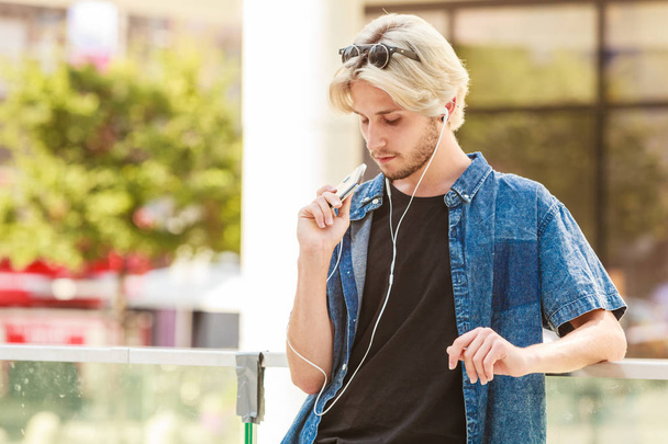 Men fashion, technology, urban style clothing concept. Hipster guy standing on city street wearing jeans outfit listening to music through earphones - 写真・画像