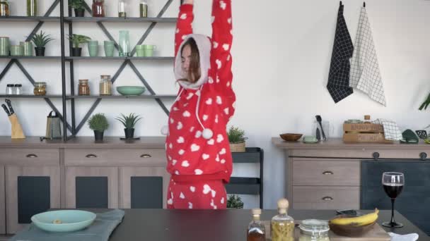 Happy ginger woman wearing hood of funny pajamas with hearts and dancing in modern kitchen behind cooking table - Imágenes, Vídeo