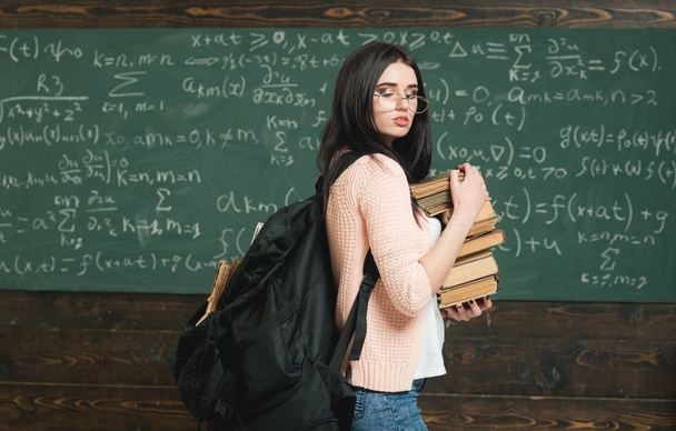 Dívka s velkým batohem drží hromadu knih, tabuli pozadí. Studentky pracuje na výzkumu. Příprava na zkoušku testu pilný student. Zavazadlo znalostí. Vynikající student hromadí znalosti - Fotografie, Obrázek