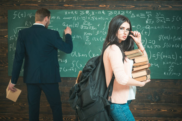 Brunette student with heap of books and heavy rucksack holding her glasses. College girl standing in front of green board while professor is writing formula - Φωτογραφία, εικόνα