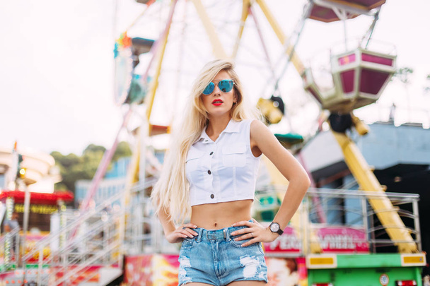 Beautiful blonde woman wearing sunglasses close-up portrait of a young sexy girl hipster in the park attraction.Stylish happy young woman wearing short denim shorts and a white T-shirt. brightred lips. - Fotó, kép