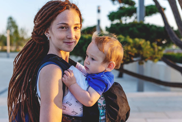 Молодая счастливая мама с маленьким сыном в рюкзаке Эрго путешествуют вместе. Солнечный летний день. Концепция современной матери
 - Фото, изображение