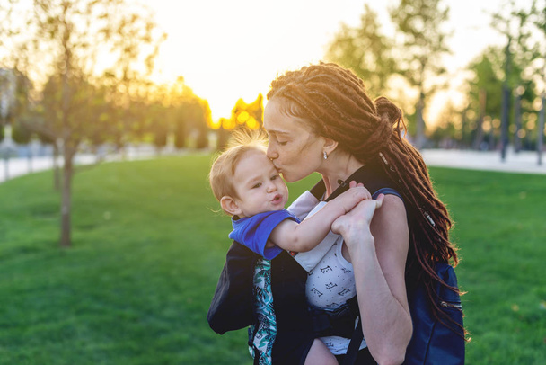 赤ちゃんの息子と若い幸せママは、公園を散歩してエルゴ バックパックします。晴れた夏の日。現代の親の概念 - 写真・画像