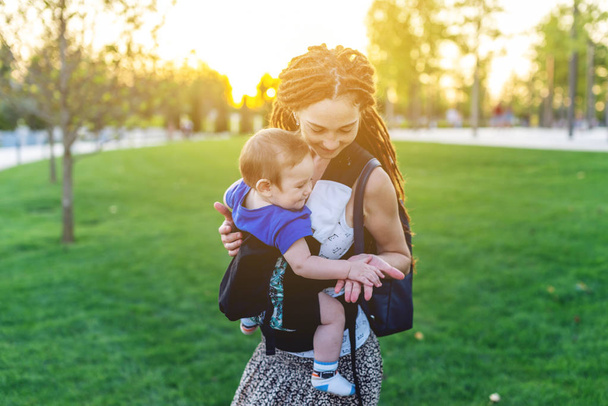 赤ちゃんの息子と若い幸せママは、公園を散歩してエルゴ バックパックします。晴れた夏の日。現代の親の概念 - 写真・画像
