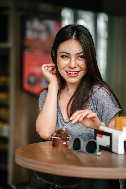 Portrait of a young Japanese Asian tourist woman smiling as she enjoys a chocolate ice cream at a table in a cafe on a hot day. - Photo, Image
