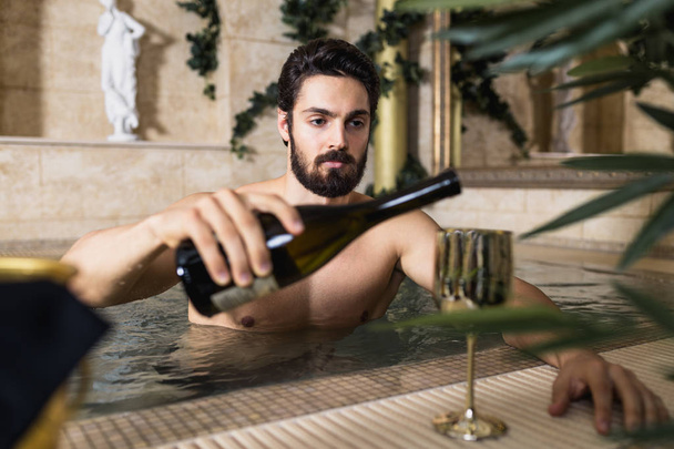 Young handsome man enjoying and relaxing in spa center. He sitting in jacuzzi and drinking wine. - Foto, Imagem