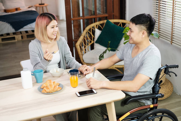 Asian couple breakfast at home.wife holding hand and encourage disabled husband sitting in wheelchair at kitchen table.couple in home concept - Photo, Image