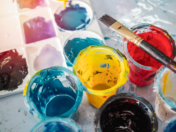 Acrylic colour in plastic cups with paintbrush and colorful artist palette on dirty paints background in art studio. - Photo, Image